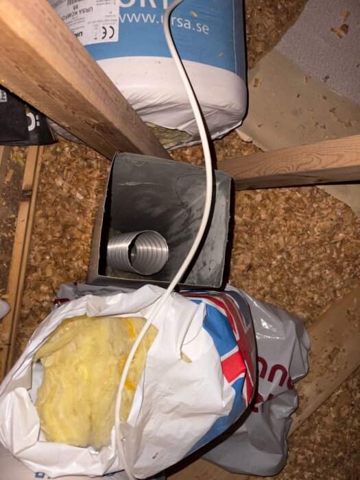 Ventilationsrör på vinden, omgivet av träreglar och isolering i plastpåsar; sågspån på golvet.