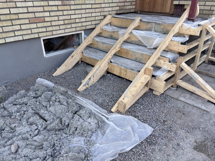 Nybyggda trappsteg i trä, täckta med plast på en gjuten yta. Byggmaterial och grus syns runt trappan under konstruktion.