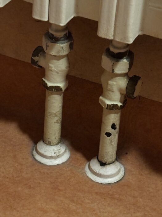 Vita rör under ett element med muttrar och möjliga avstängare, mot en brun golvyta.