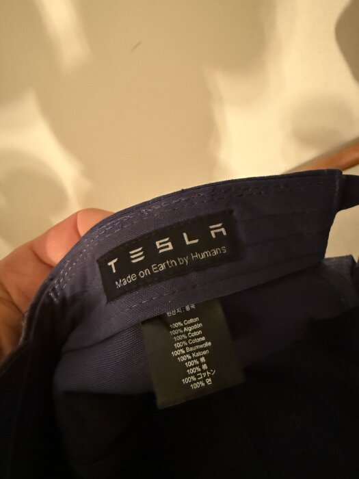 Etikett på insidan av en keps, text: "Tesla Made on Earth by Humans" samt information om bomullsinnehåll på flera språk.