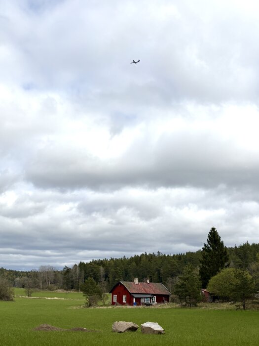 Rött hus på landet omgiven av gröna fält och skog, med ett flygplan synligt på himlen över ett molntäckt landskap.