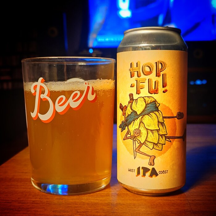 Ett glas fyllt med IPA bredvid en burk med etiketten "Hop-Fu! West Coast IPA" på ett bord.