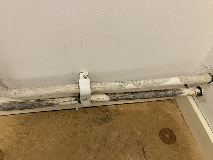 Vita och svarta rör med tecken på möjlig mögel och flagnande färg, fastsatta mot en vit vägg och brun golvyta.