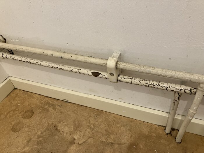 Spruckna och missfärgade rör mot en vit vägg, potentiellt angripna av mögel, placerade ovanpå trä- eller laminatgolv.