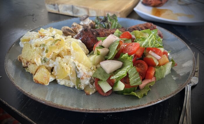 Tallrik med grillat kött, potatisgratäng och sallad med tomat, gurka, rädisa och salladslök. Serveras på en blå tallrik.