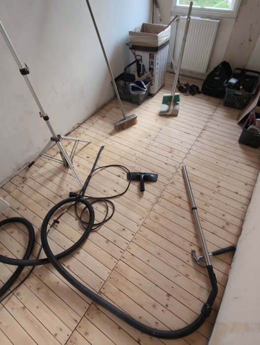 Rum under renovering med synligt råspontgolv, verktyg och städutrustning. En dammsugarslang och olika borstar ligger på golvet vid väggen.