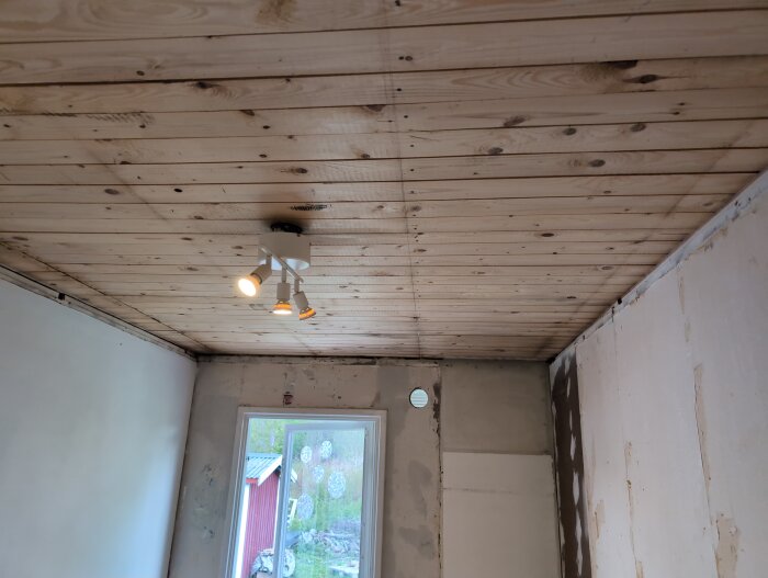 Ett rum med råsponttak och delvis renoverade väggar, ett fönster och en taklampa med flera spotlights syns i bilden.