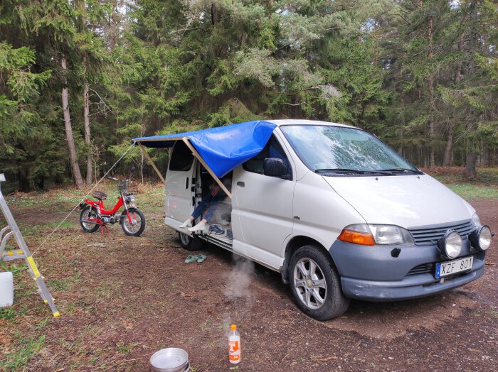 Vit skåpbil med blå markis i skogen, motorcykel bredvid, någon sitter under markisen. Flaskor på marken, det regnar lätt.