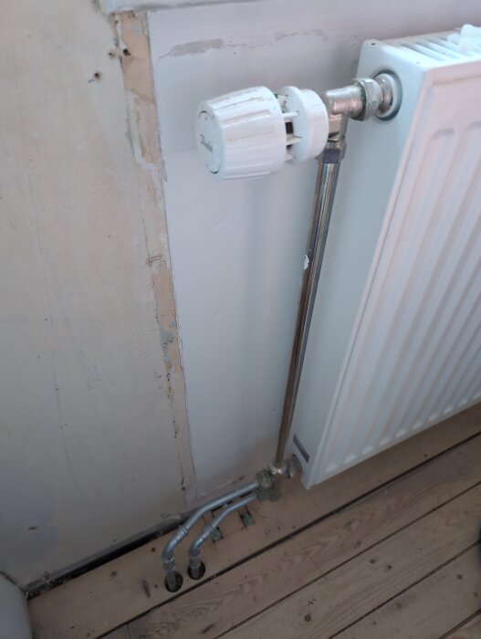 Radiator med synliga rör och termostatventil på ojämn vägg med tapetrester i renoverat rum. Trägolv nedanför.