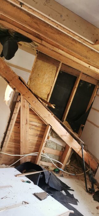 En träbalk i en byggd kupa, synlig isolering och träreglar; trasigt takmaterial och verktyg på golvet i ett renoveringsprojekt.