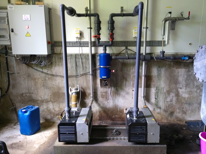 Två oanvända vakuumpumpar placerade i en verkstad med rördragning och en blå behållare i bakgrunden.