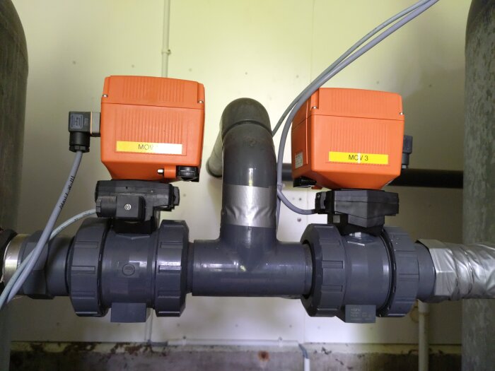 Två motorstyrda kulventiler med orangea kontrollboxar monterade på rör i en inomhusmiljö.