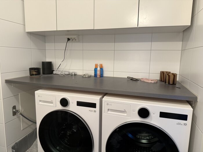 Grå bänkskiva ovanpå vit tvättmaskin med föremål som burkar, kablar och solkräm. Vita väggplattor och skåp i bakgrunden.