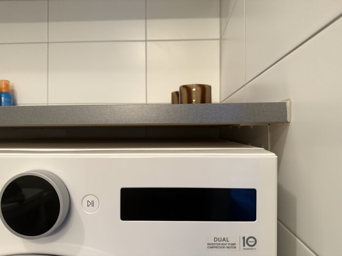 Bänkskiva i badrum monterad ovanpå tvättmaskin, med flaskor och mugg ovanpå. Vägg av vita kakelplattor syns i bakgrunden.