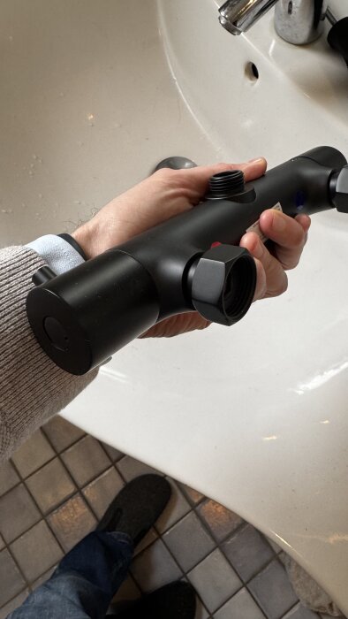 Hand håller i ett svart takduschset, synlig gängad anslutning, ovanför tvättställ med kakelgolv i bakgrunden.