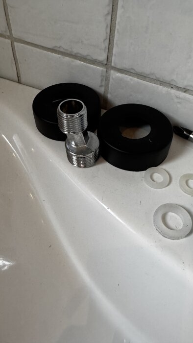 Komponenter för blandare; gängad metallhylsa, svarta och vita tätningar, liggande på vit yta intill kaklat väggparti i badrum.