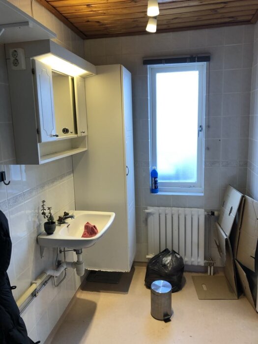 Badrum med handfat, spegelskåp, fönster och element. En papperskorg och svart soppåse på golvet, samt kartonger lutade mot väggen.