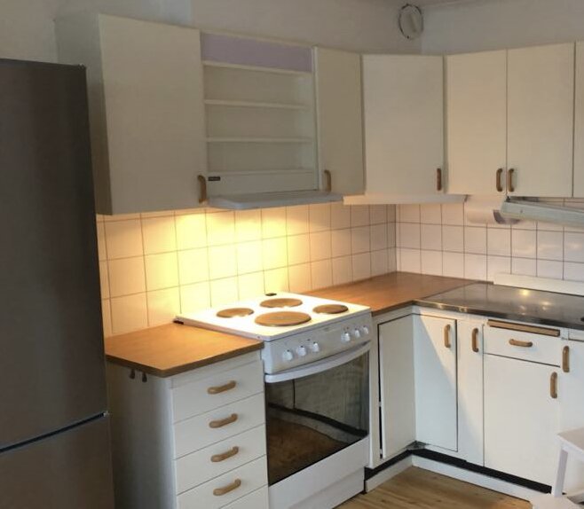 Kök med vita skåp, trähandtag, spis med fyra plattor, kylskåp, vita kakelplattor och belysning under skåpen.