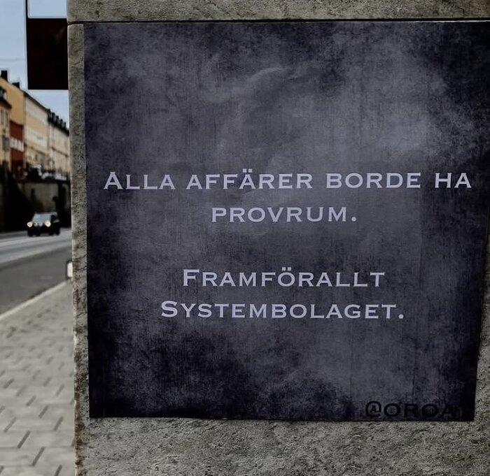 Skylt med texten "Alla affärer borde ha provrum. Framförallt Systembolaget" på en gata med bilar och byggnader i bakgrunden.