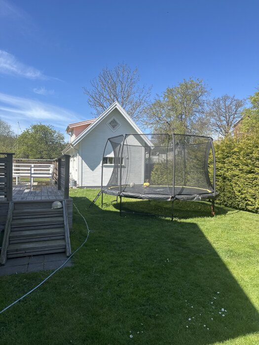 Trädgård med en studsmatta på gräsmattan bredvid en altan, träd och liten byggnad i bakgrunden under en klarblå himmel.