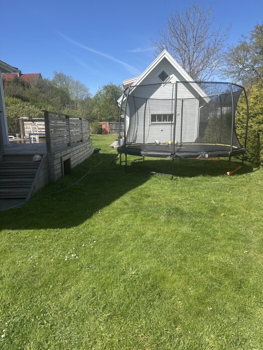 Trädgård med gräsmatta, stor studsmatta och altan. Bakom ligger ett litet vitt hus. Perfekt plats för att bygga eller sänka ner en pool.