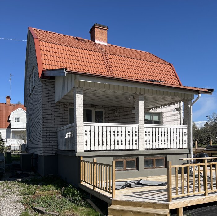 Vit tegelvilla med rött tak och altan med vita räcken, belägen vid gräsyta. Under altanen finns ett garage med små fönster.