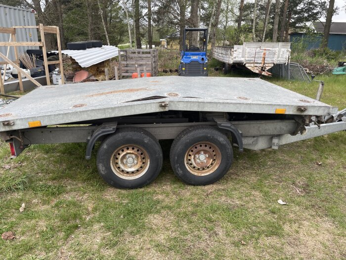 En metaltrailer med två hjul står på gräsmark, omgiven av en byggarbetsplats med material och verktyg.