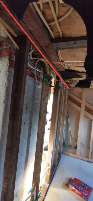 Insida av ett rum under renovering med träbjälkar, elledningar och isolering samt en röd linje över bilden som markerar en diagonal balk.