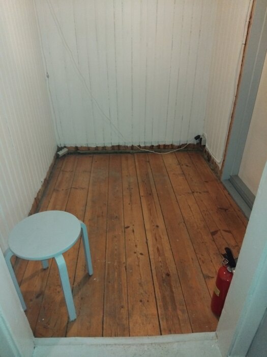 Liten hall med trägolv i en gammal stuga, träplankor som ska rätas upp och återanvändas. En pall och brandsläckare syns i hörnet.