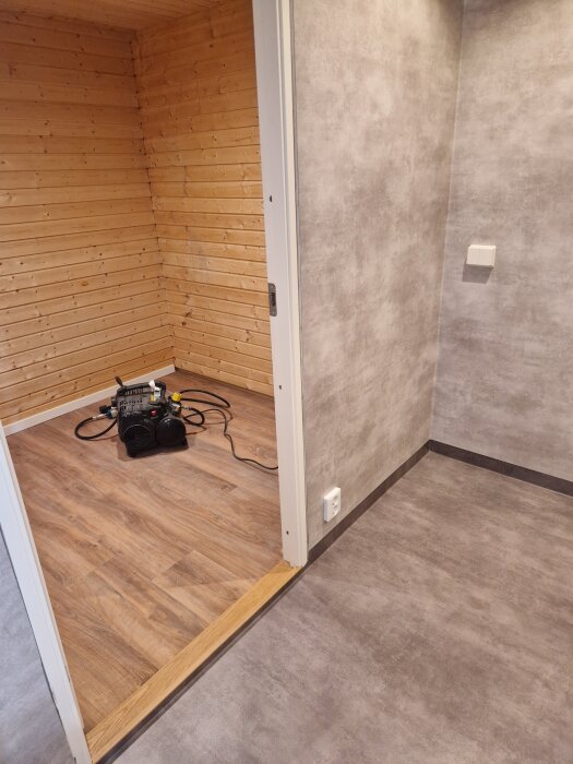 Renoverat förråd med träpaneler och LVT-golv, intill grå tvättstuga med Tarkett Aquarelle golv. Elektrisk utrustning på golvet.