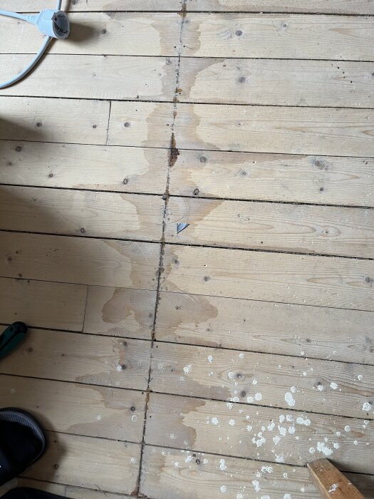 Råspontgolv med stora missfärgningar och vita fläckar, eventuellt tecken på vattenskada eller röta. En stickkontakt och verktyg syns vid kanten.