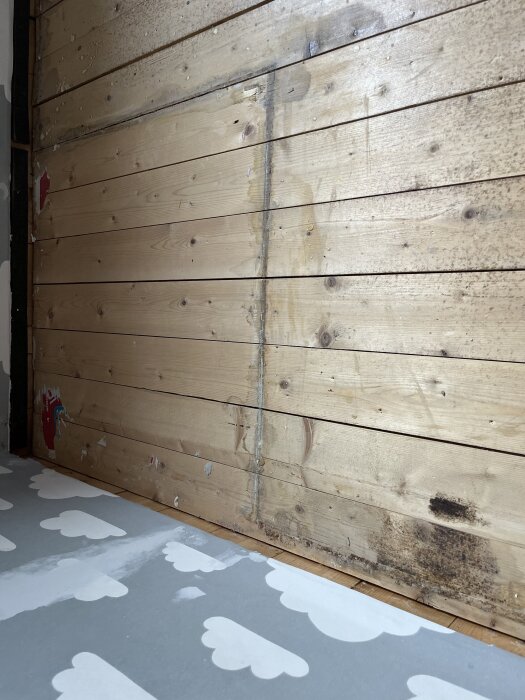 Råspontgolv med misstänkta vattenskador och missfärgningar, delvis omgiven av ett golv med molnmönster.