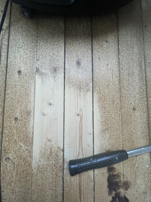 Råspontgolv med missfärgningar, potentiella vattenskador eller röta; svart handtag av verktyg syns på bilden.