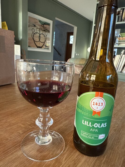 Ett glas med röd dryck bredvid en flaska Lill-Olas APA på ett träbord, i en hemtrevlig miljö med konst på väggen.