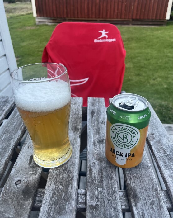 Glas med öl bredvid en burk Nya Carnegie Jack IPA på en trädgårdsbord, med en röd stol i bakgrunden.