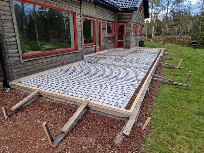 Formning av altangjutning påbörjad med trästöd och armeringsjärn framför ett trähus med röda fönsterkarmar och dörrar.