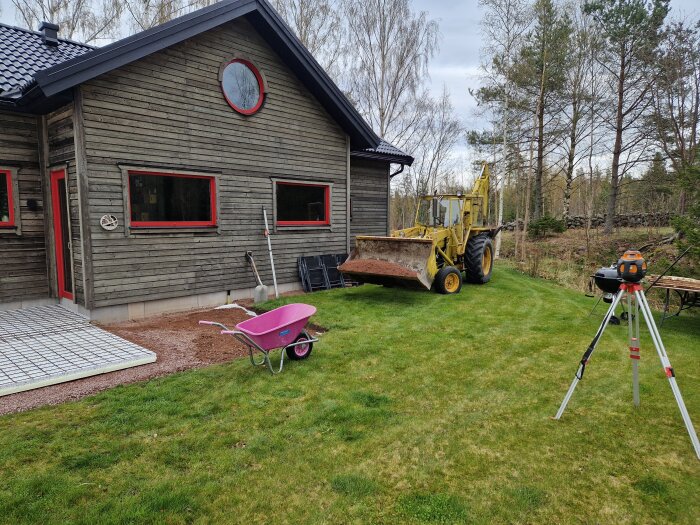 Gul traktorgrävare vid ett hus bredvid ett område förberett för altangjutning, med rosa skottkärra och utrustning på en gräsmatta.