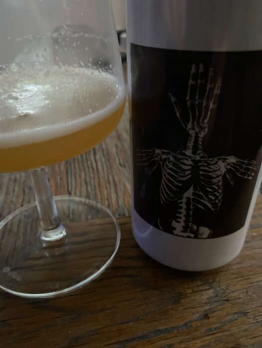 Glas med skummande trippelöl bredvid en ölburk med skelett på etiketten, stående på träbord.