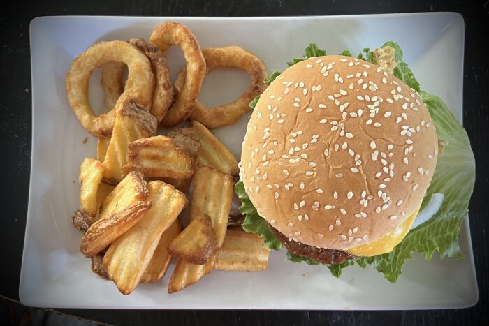 Hamburgare med sallad, ost, tomat och lök på en tallrik med lökringar och pommes frites vid sidan.