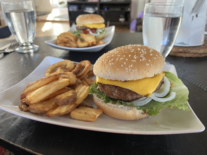 Cheeseburgare med ost, lök, sallad och dressing serverad med pommes frites och lökringar på en vit tallrik.