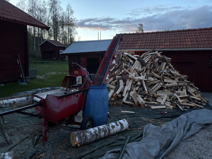 Vedklyvningsmaskin och stor vedhög på grusuppfart omgiven av röda träbyggnader, skymning i bakgrunden.