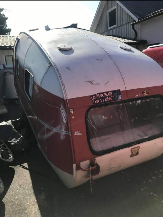 Gammal SMV10 husvagn från 1962 i dåligt skick, ståendes på en parkeringsplats framför ett hus. Röd och vit med rostiga detaljer.