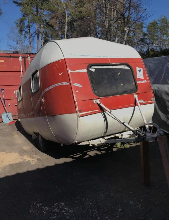 Äldre SMV husvagn, modell "Ägget" från 1962 i slitet skick, med en rödmålad sektion, synlig på en parkeringsplats med träd i bakgrunden.