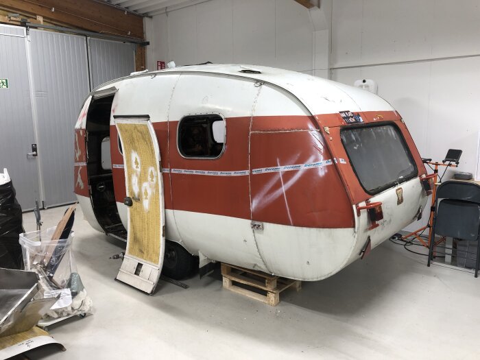 En vit och röd husvagn under renovering, med dörren borttagen och placerad vid sidan om, står på en träpall i en verkstad.