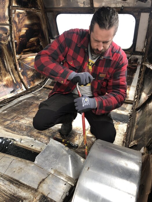 Man i rutig skjorta arbetar med att bryta upp golvplåt inne i en husvagn under renovering. Verktyg som hammare och kofot syns.