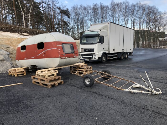 Röd och vit husvagn utan underrede står på träpallar bredvid en lastbil på asfaltsplan, underredsram ligger på marken framför.