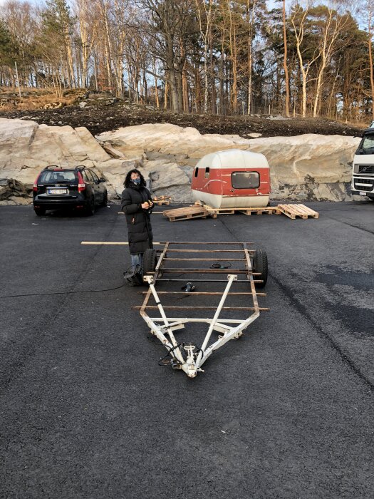 En kvinna står vid ett underrede och en fristående husvagnskropp på en asfalterad yta med skog i bakgrunden.