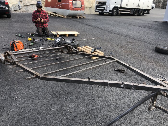 Underrede av en campingvagn på en asfaltsyta, man i röd skjorta knäböjer med verktyg, rost och krockskada synliga på ramen.