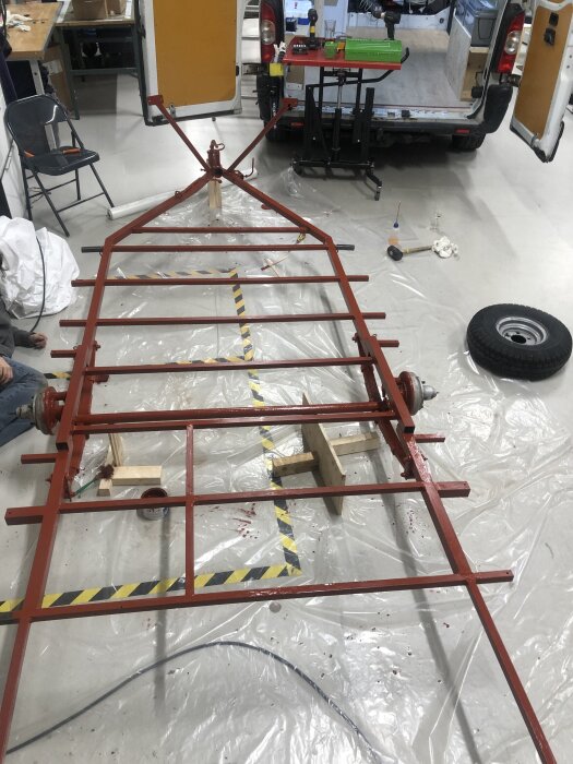 Ett renoverat underrede av en vagn med röd-primade metallramar på en verkstadsgolvet, omgiven av verktyg och plastskydd.