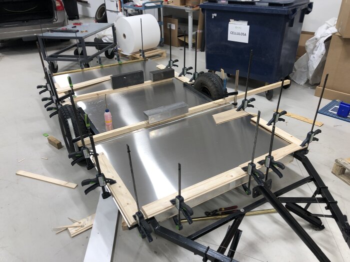 Byggprojekt med ett underrede där ny aluminiumplåt limmas och kläms fast med tvingar, verktyg och byggmaterial syns i bakgrunden.